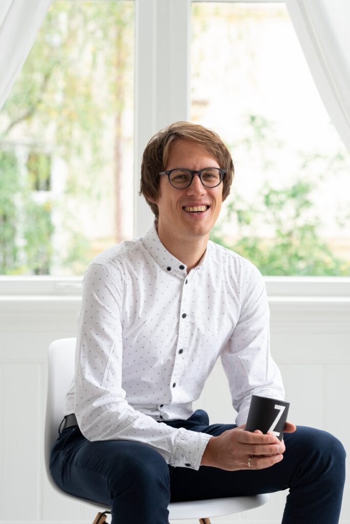 Portrait von Oliver Schöndorfer sitzend auf einem Sessel mit einer schwarzen Kaffeetasse in der Hand die ein weißes Z zeigt. Oliver lächelt freundlich in die Kamera